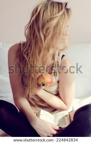 portrait of sexy blonde girl having fun happy holding book & big red apple sitting on white bed on light copy space background