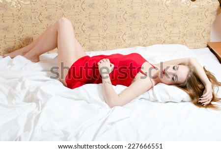 charming sleeping beauty: elegant sensual beautiful female having fun in red dress dreaming or sleeping with eyes closed lying on the white bed, copy space background portrait
