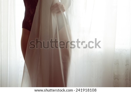 closeup on silhouette of hiding behind tulle curtain pretty lady with perfect fit body in black combies dress standing on light window copy space background