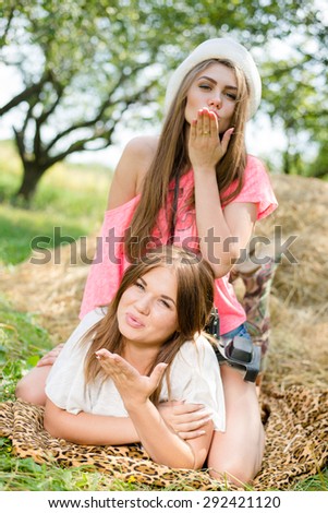 Blowing kiss 2 pretty girls best friends having fun relaxing lying on hay happy smiling & looking at camera on green summer outdoors copy space background