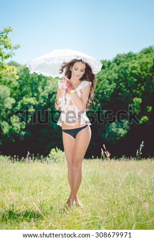 Portrait of amazing young brunette sensual girl with perfect body in lingerie standing on green grass under white umbrella relaxing on summer day outdoors background