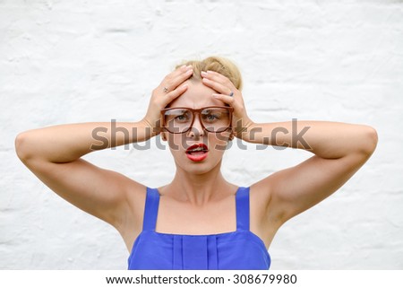 Picture of worried sad beautiful lady in glasses holding her hands on head over white brick wall background