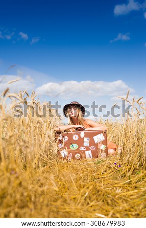 Young beautiful romantic woman in hat sitting in field of wheat with retro suitcase over blue sky on summer day outdoors background