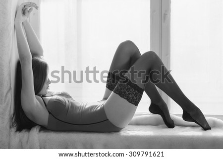 Beautiful sexy girl having fun relaxing lying on window sill on light copy space background. Black and white photography