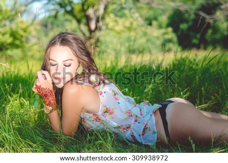 Brunette sexy girl wearing black strings lying on green grass. Woman with closed eyes and tattoo posing in flower top and gold metal watch on hand.