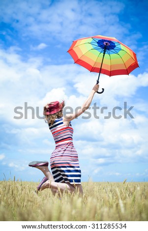 Happy female in stripped dress and red hat jumping with rainbow umbrella in the wheat field on sunny blue sky outdoors background