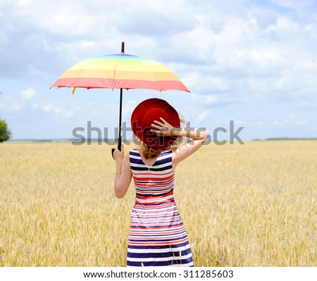 Blond woman with red hat on holding rainbow umbrella. Girl in multi-coloured stripped dress looking away in the wheat field on sunny blue sky outdoors background