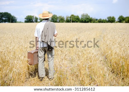 Rural man in straw hat standing in yellow wheat meadow. Man with suitcase looking away and thinhking where to go.