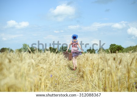 Female wearing hat with suitcase walking away through wheat field. Backview of girl in striped dress carring old suitcase over sunny blue sky background