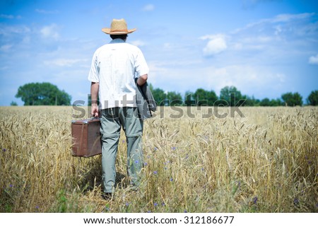 Man wearing hat with suitcase walking away through wheat field. Backview of male in white shirt carring old suitcase over blue sky outdoors background