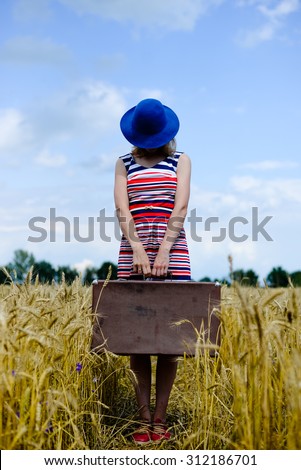 Backview of girl in striped dress carring old valize. Romantic woman wearing hat with suitcase walking away through wheat field