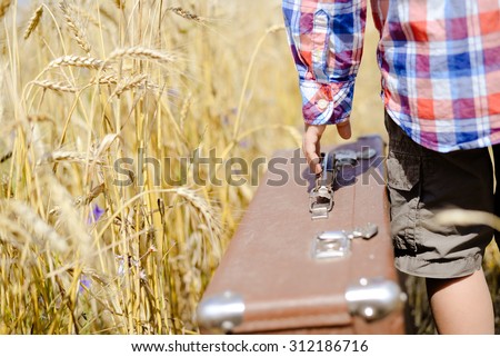 Closeup on boy's hand with big old retro leather suitcase in yellow wheat field. Closeup of hand with valize and part of shirt with shorts Imagine de stoc © 