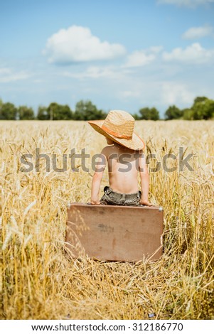 Little kid sitting on big old brown leather suitcase in summer countryside. Child with valize wearing straw hat in wheat field over sunny blue sky outdoors background Imagine de stoc © 