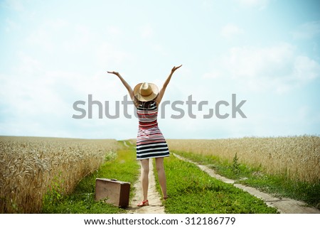 Backview of happy woman with old suitcase on countryside road. Girl with valize wearing hat and striped dress rising hands to the sunny blue sky background Imagine de stoc © 