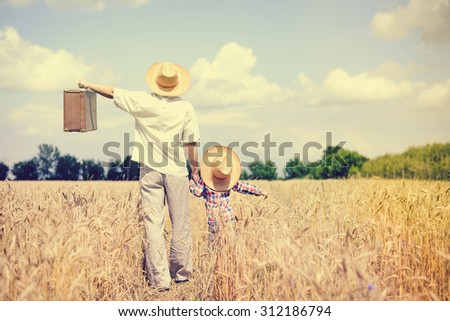 Man and boy wearing straw hats with old brown suitcase in wheat field. Happy father and son on vacation over sunny blue sky outdoors background