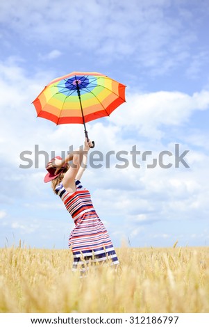 Girl rising umbrella and standing in golden wheat field. Women holding red hat to protect from wind over blue sky outdoors background
