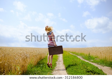 Woman with valize jumping on road between field of wheat. Backview of girl wearing hat and looking away over blue sky sunny outdoors background  Imagine de stoc © 