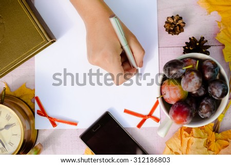 Hand writing on white paper near maple leaves and plums. Phone, clock, book and pinecones on lilac plank desk.
