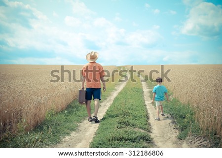 Picture of man and boy walking away on road between field of wheat over sunny blue sky background. Backview of man wearing hat with old valize and little kid. Imagine de stoc © 