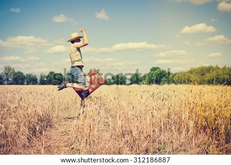 Happy jumping man wearing straw hat with valize in wheat field. He rising hand and having fun over blue sky background Imagine de stoc © 