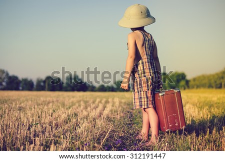 Picture of little boy wearing pith helmet and plaid romper carrying big valize in countryside . Kid in safari hat looking back on the blue sky outdoors background Imagine de stoc © 