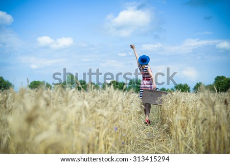 Picture of happy woman wearing blue hat with valize standing and waving hand to sky in wheat field. Backview of young girl in striped dress carring old suitcase on summer countryside background. Imagine de stoc © 