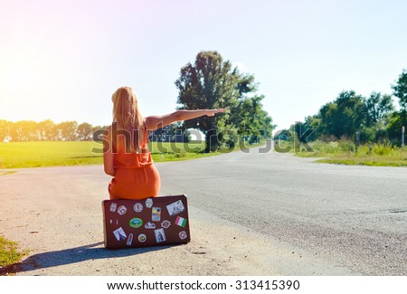 Picture of young woman sitting on suitcase on country crossroad. Backview of blond girl in orange dress hitchhiking car on sunny outdoors background. Imagine de stoc © 