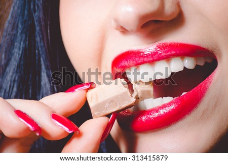 Picture closeup of beautiful woman\'s mouth biting big milk chocolate sweet. Female wearing red lipstick and has red nails.