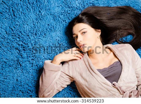 Picture of beautiful woman in beige robe lying on blue fluffy carpet. Young girl looking at camera on bright comfort indoor background.