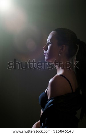 Portrait of young woman in black underwear standing with backlit. Dramatic sideview of thoughtful girl on flare blurred dark indoor background.