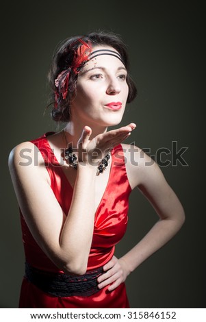 Portrait of beautiful young woman in retro dress sending air kiss. Pretty girl in 1920s styled red costume flirting on dark blurred background.