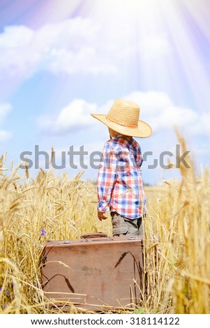 Picture of little boy wearing straw hat and plaid shirt with old suitcase. Kid standing in wheat field on blue sky sunny outdoors background.