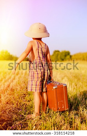 Back view picture of little boy wearing pith helmet and plaid romper carrying big brown valize case in countryside. Kid in safari hat looking back on the blue sky outdoors background Imagine de stoc © 