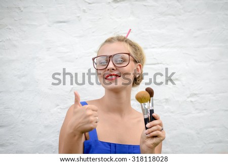 Portrait of blond girl in hipster glasses thumbing. Young woman holding makeup brushes and happy smiling on white wall background.