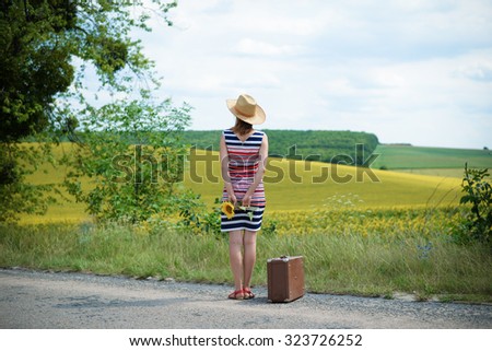 Back view of young lady standing on the road and looking away in the distance. Girl in stripped dress with straw hat on holding a sunflower behind her back. Suitcase is near by her. Imagine de stoc © 