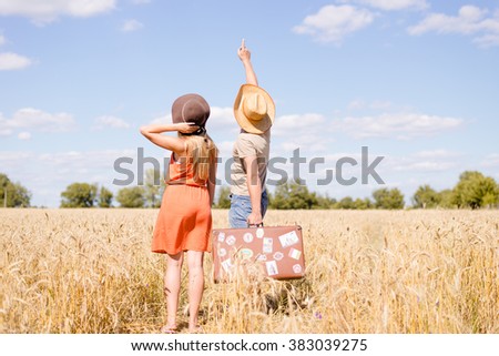 Joyful young couple having fun in wheat field. Excited man and woman pointing up holding retro leather suitcase on blue sky outdoor background. Imagine de stoc © 