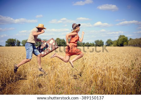 Joyful young couple having fun in wheat field. Excited man and woman jumping with retro leather suitcase on blue sky outdoor background. Imagine de stoc © 
