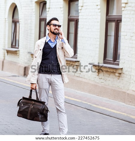 Young urban businessman on smart phone running in street talking on smartphone smiling wearing jacket and leather bag