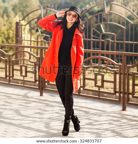 Beautiful young brunette woman in sunglasses and a hat walks through the city in the autumn sunny day. Lady dressed in a red coat, black jeans and boots