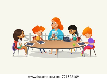 Redhead female teacher showing picture to children sitting around round table at class with laptop and tablet pc. Kids using gadgets during lesson at primary school. Colorful vector illustration.