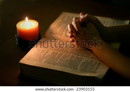 Praying hands on top of open bible, lit only by candle light
