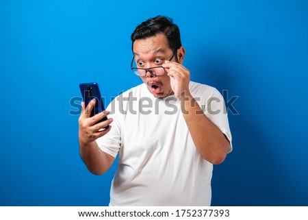 Fat Asian guy wearing a white t-shirt looks surprised at the good news he received from his smartphone. Men show shocked movements with bulging eyes while rolling down his glasses on a smartphone 商業照片 © 