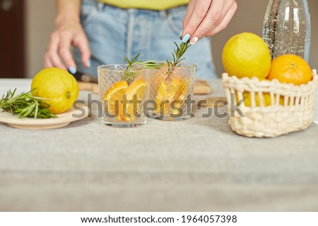 Female hand put rosemary in glasses, Woman preparing, making citrus and rosemary fresh lemonade in glass on a white table at home, summer drink, detox healthy water.