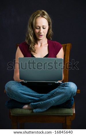 A pretty young woman works on a laptop computer while sitting cross-legged in an armchair.