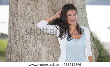Pretty young woman (20s) enjoying and looking at camera in nature. Head and shoulders with one hand behind head.