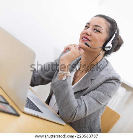 Portrait of a smiling well-dressed saleswoman with headset on in office behind her desk and laptop computer