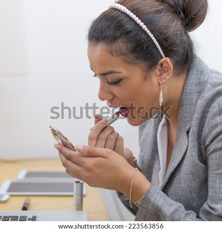 Before a meeting or before to leave the office, a beautiful brown young woman applying lipstick with little mirror at her desk.