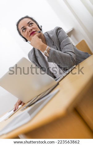 Beautiful brown young woman in office enjoying working with headset on. She is smiling behind her laptop computer on wood desk.