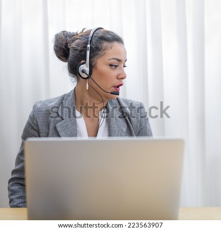 Portrait of sales woman in action sitting with headset on at her wood desk behind display computer
