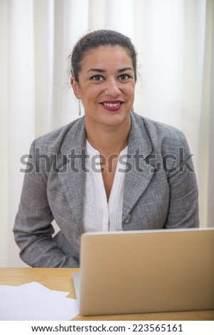 Portrait in office of businesswoman head and shoulders sitting behind her laptop computer on wood desk. She is smiling.